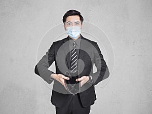 Young businessman wearing a mask, went bankrupt during COVID-19, holding empty wallet and standing in front of the gray wall