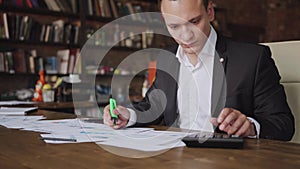 Young businessman wearing suit working with documents and calculator sitting at office desk, financial analyst using or