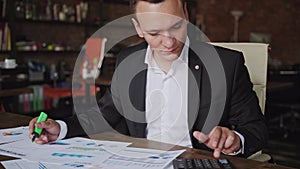 Young businessman wearing suit working with documents and calculator sitting at office desk, financial analyst using or