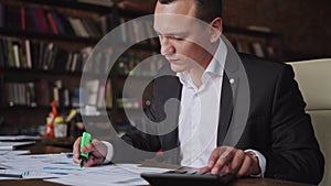 Young businessman wearing suit working with documents and calculator sitting at office desk, financial analyst using or