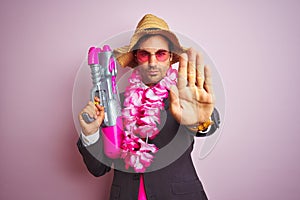 Young businessman wearing water gun hawaiian lei hat glasses over isolated pink background with open hand doing stop sign with