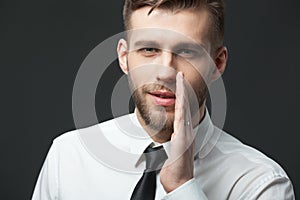 Young businessman whispering trade secrets on dark gray backgro