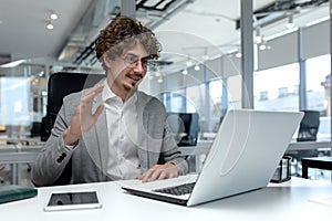 A young businessman works in the office, sits at a desk and talks on a video call through a laptop. greets and waves at