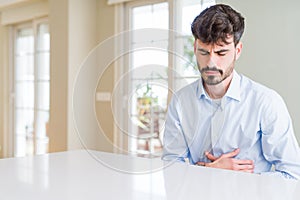 Young businesss man sitting on white table with hand on stomach because nausea, painful disease feeling unwell