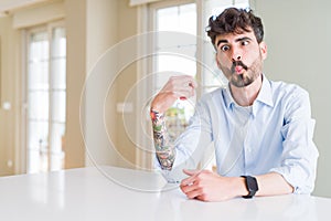 Young businesss man sitting on white table making fish face with lips, crazy and comical gesture