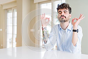 Young businesss man sitting on white table relax and smiling with eyes closed doing meditation gesture with fingers