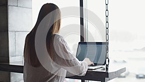 Young businesswoman dressed in white shirt is sitting in cafe at wooden table in front of laptop and talking on cell