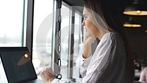 Young businesswoman dressed in white shirt is sitting in cafe at wooden table in front of laptop and talking on cell