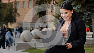 Young businesswoman freelancer student sitting outdoors reading email on laptop smiling laughing good news feels happy