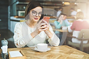 Young businesswoman in glasses and white sweater is sitting in cafe at table and using smartphone, working.E-learning