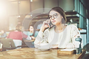 Young businesswoman in glasses and white sweater is sitting in cafe at wooden table and talking on cell phone.