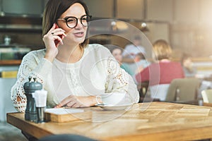 Young businesswoman in glasses and white sweater is sitting in cafe at wooden table and talking on cell phone