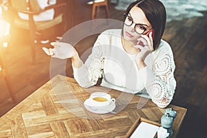 Young businesswoman in glasses and white sweater is sitting in cafe at wooden table and talking on cell phone.