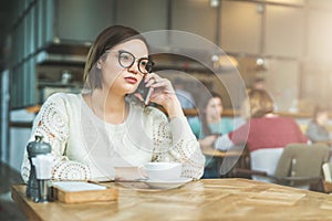 Young businesswoman in glasses and white sweater is sitting in cafe at wooden table and talking on cell phone.