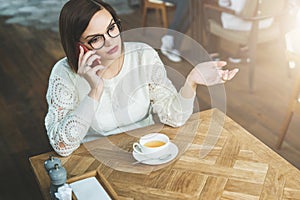 Young businesswoman in glasses and white sweater is sitting in cafe at wooden table and talking on cell phone.