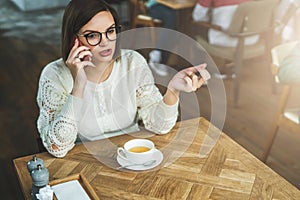 Young businesswoman in glasses and white sweater is sitting in cafe at wooden table and talking on mobile phone