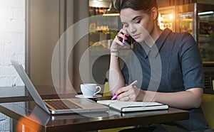 Young businesswoman in gray dress sitting at table in cafe, talking on cell phone while taking notes in notebook.