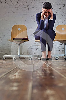 Young businesswoman with headache sitting against white brick wall at office lobby