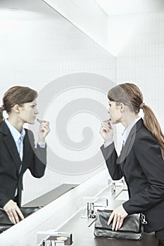 Young businesswoman with long hair looking into the mirror and putting on lipstick in the bathroom