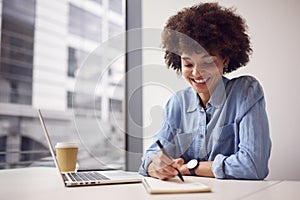 Young Businesswoman In Modern Office Working On Laptop Making Notes In Notebook