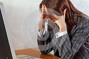 Young businesswoman or office worker in classic striped suit sits in front of the computer holding her head with hands.