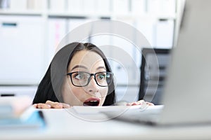 Young businesswoman peering over her desk in office in wide eyed horror or amazement