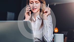 Young businesswoman in shirt is sitting in office at table in front of computer, talking on cell phone
