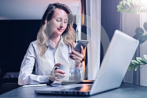 Young businesswoman in shirt is sitting in office at table in front of computer, using smartphone, looks at phone screen