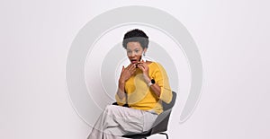 Young businesswoman with shock expression speaking on cellphone on chair against white background
