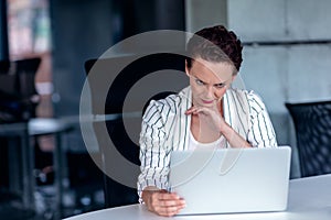 A young businesswoman sits behind her laptop computer frowning with her head in her hands because of a problem at work