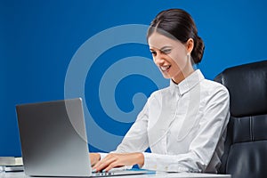 Young businesswoman sitting at her desk fiercely typing her ideas, isolated on blue background