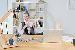 Young businesswoman sitting in office at her desk, holding a cup