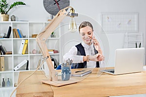 Young businesswoman sitting in office at her desk looking at wristwatch and talking