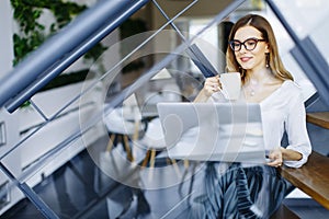 Young businesswoman sitting oh the office stairs and having coff