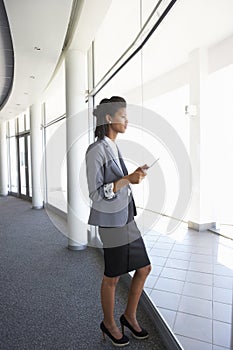 Young Businesswoman Standing In Corridor Of Modern Office Building Using Tablet Computer