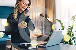 Young businesswoman is standing indoor near table in front of computer, while talking on cell phone and holding apple.