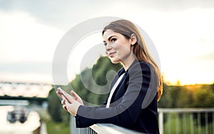 A young businesswoman standing on the river bank, using smartphone.