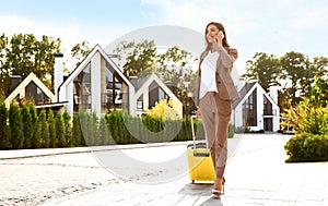 Young businesswoman with suitcase talking on phone outdoors. Moving day