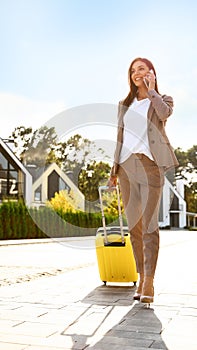Young businesswoman with suitcase talking on phone outdoors. Moving day