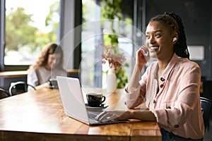 Young Businesswoman Taking Call On Wireless Earbuds Working On Laptop In Coffee Shop Or Office