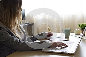 Young businesswoman teleworking sitting at a desk writing in papers and typing on her laptop in a home office. Telecommuting