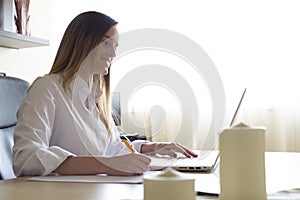 Young businesswoman teleworking sitting at a desk writing and typing on her laptop in a home office. Telecommuting and teleworking