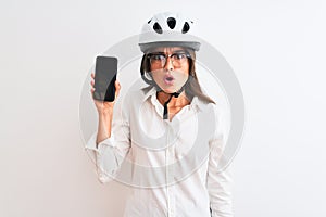 Young businesswoman wearing bike helmet holding smartphone over isolated white background scared in shock with a surprise face,