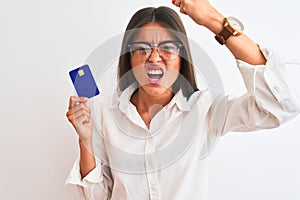 Young businesswoman wearing glasses holding credit card over isolated white background annoyed and frustrated shouting with anger,