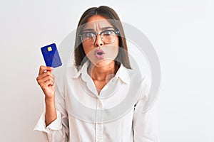 Young businesswoman wearing glasses holding credit card over isolated white background scared in shock with a surprise face,