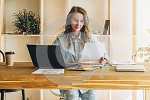 Young businesswoman woman is sitting at kitchen table, reading documents,uses laptop,working, studying. photo