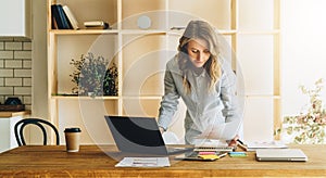 Young businesswoman woman is standing near kitchen table,reading documents,uses laptop,working, studying.