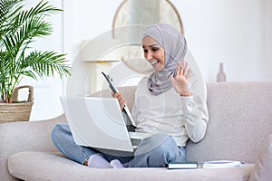 Young businesswoman working from home on laptop, sitting on sofa in lotus position and talking on video call, waving at