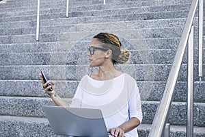 Young businesswoman working on laptop while sitting on staircases outdoors. Caucasian womantext messaging on mobile phone while