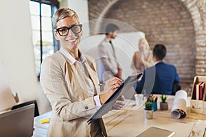 Young businesswoman writing on clipboard with team discussing project in the background